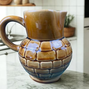 Peut inclure: Une cruche en céramique marron avec une base arrondie texturée en forme de briques et un dessus lisse et brillant. La cruche a une anse incurvée et un bec verseur. Le dégradé de couleurs passe du marron clair au marron plus foncé.