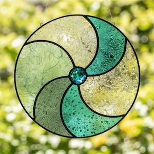 Puede incluir: Un atrapasoles redondo de vidriera con un diseño en espiral en tonos verdes y amarillos. El vidrio está segmentado con líneas negras y presenta una gema azul central. Se suspende con un alambre fino.