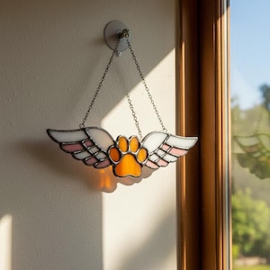 May include: Stained glass pet memorial featuring a paw print with angel wings. The wings are white and pink, and the paw print is orange. The suncatcher hangs from a chain and is attached to a wall with a suction cup.