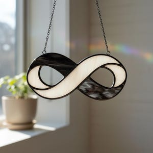 Puede incluir: Un símbolo de infinito de vidriera en blanco y negro colgado de una cadena. El símbolo está dividido en dos bucles, uno negro y otro blanco. El vidrio tiene una apariencia texturizada. La pieza está suspendida frente a una ventana.
