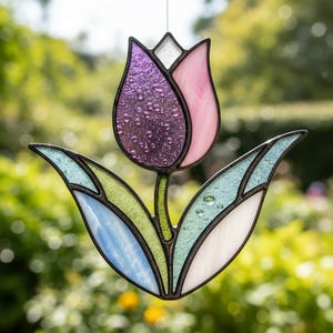 Peut inclure: Un capteur de soleil en forme de tulipe en vitrail. La fleur est violette et rose, avec des feuilles vertes et bleues. Le verre a un aspect texturé et bulleux. La tulipe est soulignée d'un métal foncé.