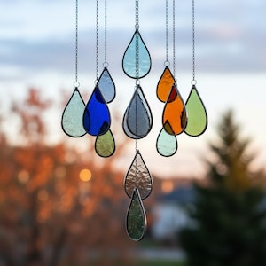 Puede incluir: Una colección de adornos de lágrima de vidrieras de colores colgando de cadenas plateadas. Las piezas de vidrio son de varios colores, incluyendo azul, verde, naranja y transparente, con un borde negro. El fondo está borroso, con follaje otoñal y un cielo suave.