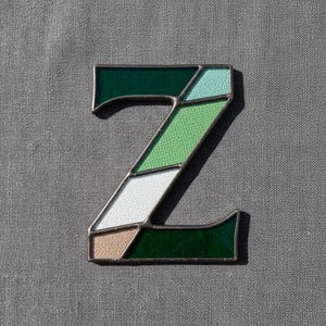 May include: A stained glass letter "Z" with a dark green border. The letter is composed of various shades of green, white, and tan glass pieces. The background is a solid gray fabric.