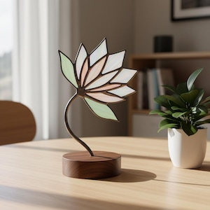 Puede incluir: Una escultura de flor de loto de vidrieras con un tallo de metal marrón y una base de madera. Los pétalos de la flor son de colores pastel blanco, rosa y verde. La escultura está sobre una mesa de madera marrón claro.