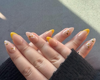 May include: Almond-shaped artificial nails with a yellow French tip design. The nails feature a clear base with small white daisy and bee decals. The hands are resting on a dark gray surface, with a black sleeve visible.