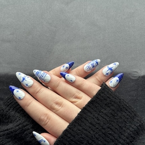May include: Almond-shaped artificial nails with a white base and blue designs. The designs include starbursts, swirls, and text. The nails are on a hand wearing a black sweater.