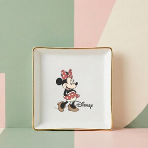 May include: Square white ceramic trinket dish with gold trim featuring a Minnie Mouse graphic. The cartoon character is wearing a red polka dot bow and skirt. The Disney logo is printed below the character.