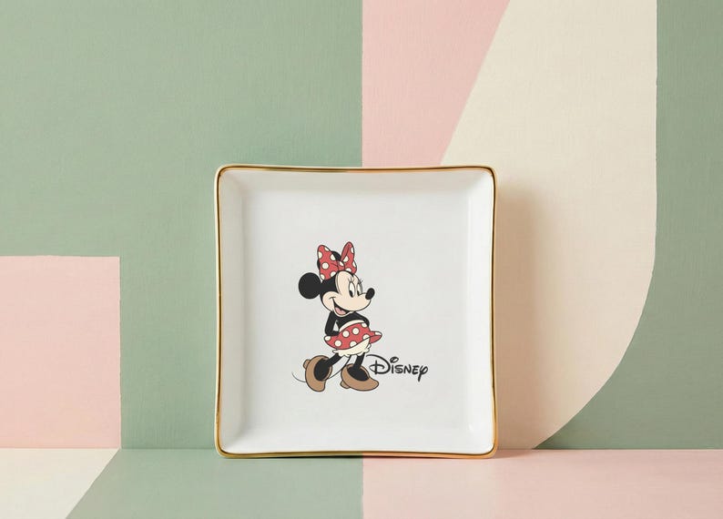 May include: Square white ceramic trinket dish with gold trim featuring a Minnie Mouse graphic. The cartoon character is wearing a red polka dot bow and skirt. The Disney logo is printed below the character.