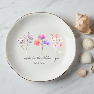 May include: A white ceramic plate with a gold rim, featuring watercolor flowers and the text "consider how the wildflowers grow, Luke 12:27." Several seashells are arranged around the plate on a marble surface.