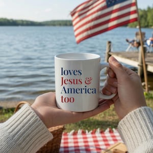 May include: White ceramic mug with the text "loves Jesus & America too" in red and blue. The mug is held in front of a lake with an American flag waving in the background. A picnic basket and red and white checkered blanket are in the foreground.