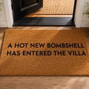 May include: A rectangular doormat made of natural fibers, with the phrase "A HOT NEW BOMBSHELL HAS ENTERED THE VILLA" printed in black. The mat is placed in front of a doorway, with potted plants on either side.