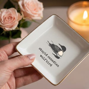 May include: A square, white ceramic trinket dish with gold trim, featuring a black and white illustration of a bird and the text "stupid canadian wolfbird." The dish is held in a hand, with a lit candle and pink roses in the background.