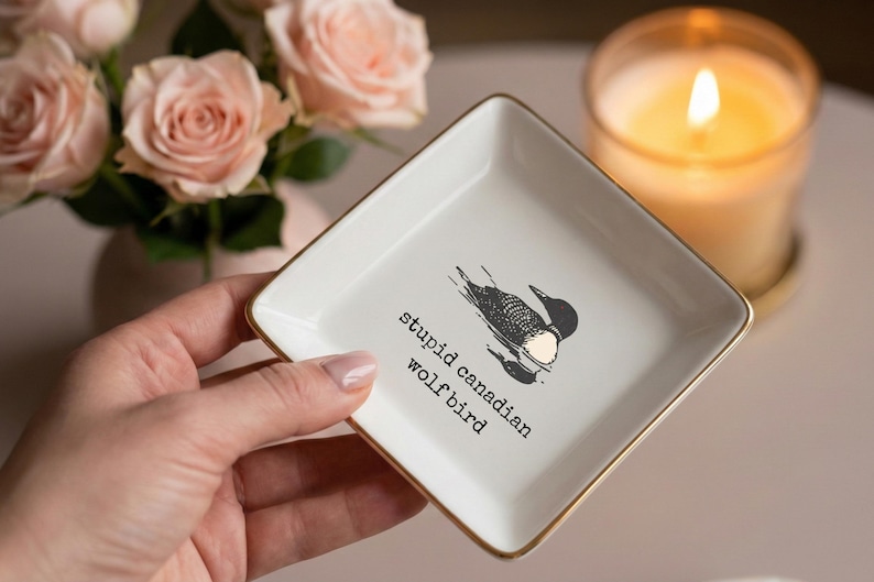 May include: A square, white ceramic trinket dish with gold edging, displaying a black and white bird illustration and the text "stupid canadian wolfbird." The dish is held in a hand, with a lit candle and pink roses in the background.