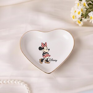 May include: A heart-shaped white ceramic trinket dish with a gold rim, featuring a Minnie Mouse illustration. The dish has the Disney logo and is next to two gold rings and a bouquet of white flowers.