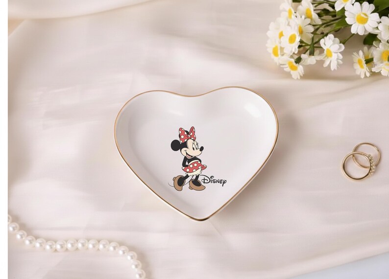 May include: A heart-shaped white ceramic trinket dish with a gold rim, featuring a Minnie Mouse illustration. The dish has the Disney logo and is next to two gold rings and a bouquet of white flowers.