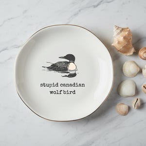 May include: White ceramic plate with a gold rim, featuring a black and white illustration of a loon and the text "stupid canadian wolf bird." Several seashells are arranged around the plate.