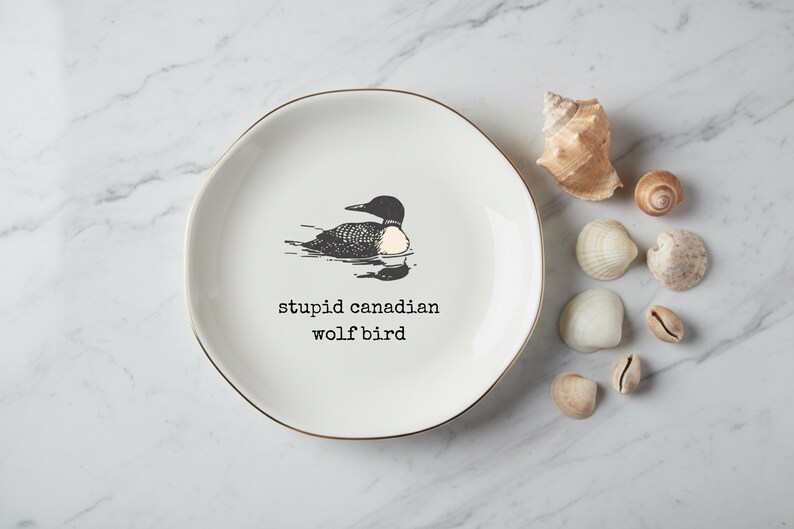 May include: White ceramic plate with a gold rim, featuring a black and white illustration of a loon and the text "stupid canadian wolf bird." Several seashells are arranged around the plate.