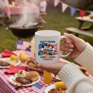 May include: White ceramic mug featuring Mickey and Minnie Mouse with the text "250th Years Anniversary America" and the USA flag. The mug is held in front of a picnic scene with food, flags, and a grill.