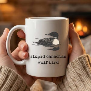 May include: White ceramic mug featuring a black and white illustration of a loon on water. The mug has the text "stupid canadian wolf bird" printed in black. The mug has a curved handle.