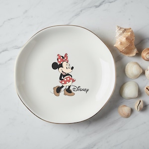 May include: A white ceramic plate with a gold rim featuring a Minnie Mouse graphic. Minnie is wearing a red polka dot bow and dress. The Disney logo is printed below. Seashells are arranged around the plate.