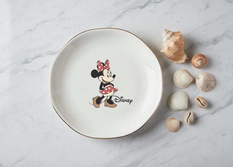 May include: A white ceramic plate with a gold rim featuring a Minnie Mouse graphic. Minnie is wearing a red polka dot bow and dress. The Disney logo is printed below. Seashells are arranged around the plate.