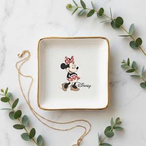 May include: A square, white ceramic trinket dish with gold trim, featuring a Minnie Mouse graphic. The dish is surrounded by gold necklaces and sprigs of greenery, set against a marble background. The Disney logo is printed on the dish.
