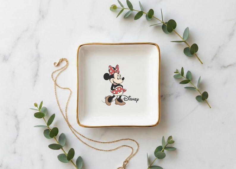May include: A square, white ceramic trinket dish with gold trim, featuring a Minnie Mouse graphic. The dish is surrounded by gold necklaces and sprigs of greenery, set against a marble background. The Disney logo is printed on the dish.