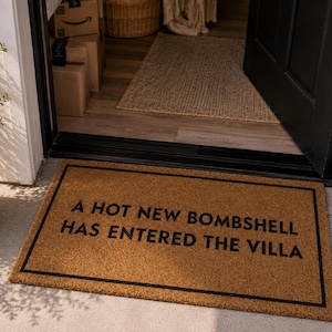 May include: A rectangular doormat with a black border and the text "A HOT NEW BOMBSHELL HAS ENTERED THE VILLA" in black. The mat is made of a light brown material and is placed in front of a doorway.