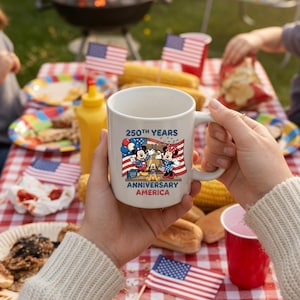 May include: White ceramic mug featuring Mickey and Minnie Mouse with the text "250th Years Anniversary America" and an American flag design. The mug is held in front of a picnic scene with food and American flags.