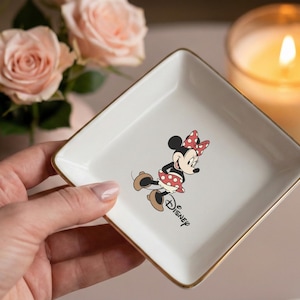 May include: A white square trinket dish with gold trim featuring a Minnie Mouse graphic. Minnie Mouse is depicted in her classic outfit with a red polka dot bow and dress. The word "Disney" is printed below the character.