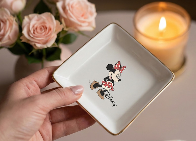 May include: A white square trinket dish with gold trim featuring a Minnie Mouse graphic. Minnie Mouse is depicted in her classic outfit with a red polka dot bow and dress. The word "Disney" is printed below the character.