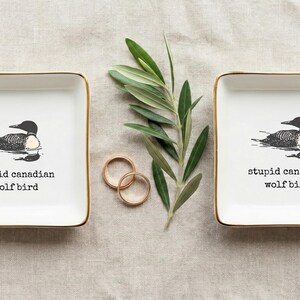 May include: Two square white ceramic ring dishes with gold trim, each featuring a black and white illustration of a loon and the text "stupid canadian wolf bird". Two gold rings and a sprig of greenery are also present.