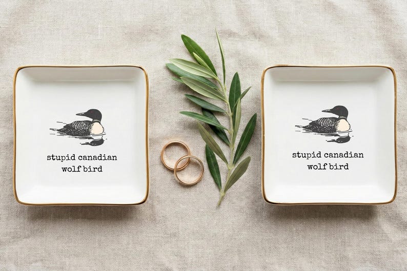 May include: Two square white ceramic ring dishes with gold trim, each featuring a black and white illustration of a loon and the text "stupid canadian wolf bird". Two gold rings and a sprig of greenery are also present.