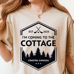 May include: Beige t-shirt with a black and white graphic. The graphic reads "I'M COMING TO THE COTTAGE" with crossed golf clubs, the year 2025, and a forest scene. The text "COASTAL CANADA" is also included.