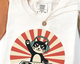 May include: Cream-colored t-shirt featuring a black cat DJ with headphones, a turntable, and a red and beige sunburst design. The shirt has the Comfort Colors brand tag. A gold necklace and a brown hat are also visible.