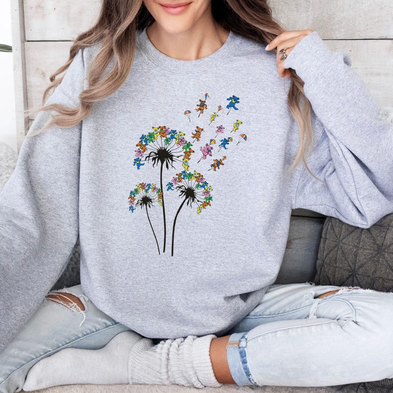 Puede incluir: Una sudadera gris claro con un dise&ntilde;o de diente de le&oacute;n colorido. Las cabezas de diente de le&oacute;n est&aacute;n compuestas por osos bailarines en varios colores, con algunos osos que parecen flotar con peque&ntilde;os paraca&iacute;das. La sudadera es de estilo casual y c&oacute;modo.