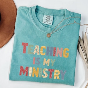 May include: A turquoise Comfort Colors t-shirt with the phrase "TEACHING IS MY MINISTRY" in colorful, patchwork-style letters. A gold necklace with a pendant rests on the shirt. The shirt is size S-P-CH.