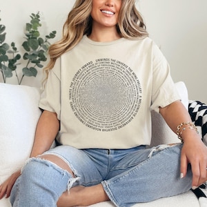 Puede incluir: Camiseta beige con un diseño circular de texto. El texto está dispuesto en círculos concéntricos, con frases como "Unwinds the Universe" y "The Journey Inward". Una prenda casual para el día a día.