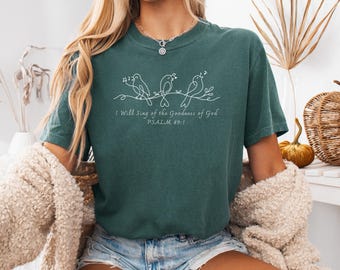 May include: A teal t-shirt featuring a white illustration of three birds singing on a branch. The text "I Will Sing of the Goodness of God, Psalm 89:1" is printed below the birds. The shirt has a crew neck and short sleeves.