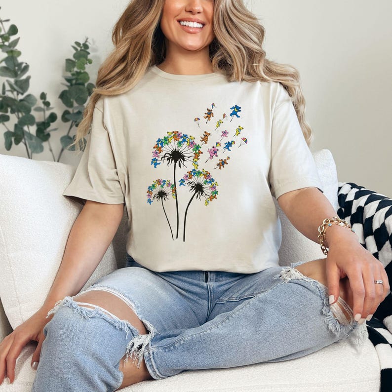 Puede incluir: Camiseta beige con un dise&ntilde;o de diente de le&oacute;n y osos bailarines de colores. El gr&aacute;fico muestra tres tallos de diente de le&oacute;n negros con cabezas de diente de le&oacute;n llenas de osos de colores. Los osos son de varios colores, incluyendo azul, amarillo, verde y rosa.