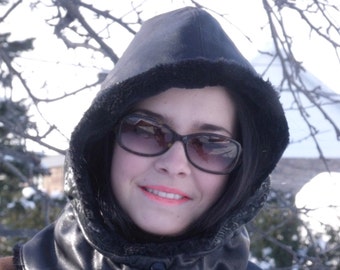 May include: A woman wearing a black leather hooded jacket with a fur trim. The jacket has a high collar and a drawstring closure. The woman is wearing sunglasses and has a neutral expression.