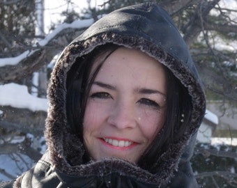 May include: A woman wearing a black hooded jacket with a fur trim. The jacket has a zipper closure and a large collar. The woman is smiling and looking at the camera. The background is a snowy landscape.
