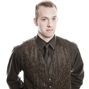 May include: A man wearing a black button-up shirt with a black and gold patterned waistcoat and a black tie.