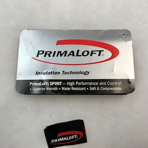 以下が含まれることがあります： 「PRIMALOFT」の文字が大きく太字で書かれた、シルバーと黒のタグ。タグには「Insulation Technology」と「PrimaLoft SPORT - High Performance and Comfort」の文字も含まれています。また、「PRIMALOFT」のロゴが入った小さな黒いタグも表示されています。