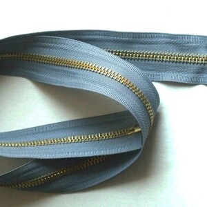 May include: A long, gray zipper with a brass slider and pull tab. The zipper is coiled and ready to be sewn into a garment.