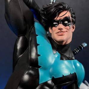 Peut inclure: Une figurine détaillée de Nightwing portant un costume noir à la finition texturée avec un emblème d'oiseau bleu vif sur la poitrine. Le personnage porte un masque noir, a des cheveux foncés et porte deux bâtons escrima dans le dos.