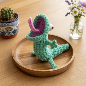 May include: A crocheted lizard figurine in light green with a wide-open mouth, a purple interior, and a protruding pink tongue. The creature has a single visible white eye with a dark pupil, small limbs, and a textured body made of yarn stitches.