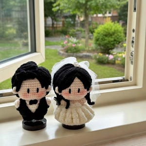 Könnte beinhalten: Zwei gehäkelte Puppen, die ein Paar in Hochzeitskleidung darstellen. Eine Puppe trägt einen schwarzen Anzug mit Fliege, die andere ein weißes Kleid mit einem transparenten Schleier. Beide haben schwarzes Haar, helle Haut und rosa Wangen.
