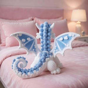 May include: A handcrafted crochet dragon plush toy featuring a white body with light blue accents on the wings, a curled tail, and a textured spine made of small, round, stitched spheres. The dragon is depicted from a rear-facing perspective.