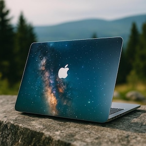 Könnte beinhalten: Ein Laptop mit dunkelblauem Deckel, der ein Sternengalaxie-Design zeigt. Das Apple-Logo ist auf dem Deckel sichtbar. Der Laptop ist geöffnet und liegt auf einer Steinoberfläche, mit einem verschwommenen natürlichen Hintergrund.